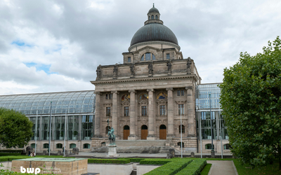 Das Bild zeigt ein historisches Gebäude mit einer großen Kuppel und Säulen an der Vorderseite. Vor dem Gebäude steht eine Statue eines Reiters. Im Hintergrund ist ein modernes Gewächshaus sichtbar, umgeben von gepflegten Gartenanlagen. Der Himmel ist bewölkt.