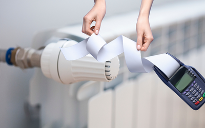 Eine Hand hält ein Stück Papier, das von einem Heizkörper herab hängt, während die andere Hand ein digitales Thermostat zeigt. Die Darstellung vermittelt einen Bezug zur Heizungssteuerung und Ablesung von Heizdaten.