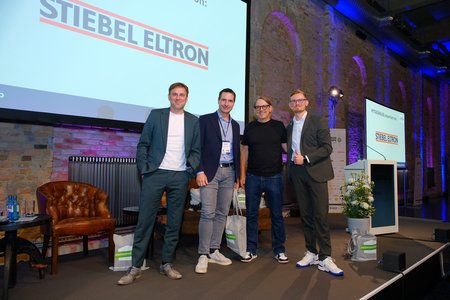 Vier Personen stehen lächelnd auf einer Bühne vor einem Bildschirm mit der Aufschrift "STIEBEL ELTRON". Sie tragen verschiedene Business-Outfits und wirken entspannt. Im Hintergrund sind ein paar Möbel und Pflanzen zu sehen, die eine freundliche Atmosphäre schaffen.