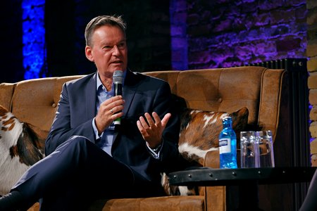 Ein Mann in einem Anzug sitzt auf einem braunen Sofa und spricht mit einem Mikrofon in der Hand. Er ist in einem Gespräch und gestikuliert mit seiner anderen Hand. Im Hintergrund sind farbige Lichter zu sehen. Auf einem Tisch neben ihm stehen Wasserflaschen.