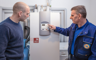 Ein Techniker erklärt einem Mann die Bedienung eines Heizungsgeräts. Der Techniker zeigt auf das Display des Geräts, während der Mann aufmerksam zusieht. Im Hintergrund sind Rohre sichtbar.