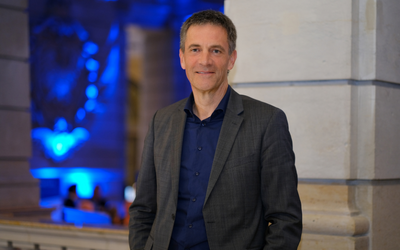 Ein Mann in einem grauen Anzug und blauen Hemd steht lächelnd an einer Säule. Im Hintergrund sind schwaches bläuliches Licht und weitere Personen sichtbar, die sich in einem eleganten Raum aufhalten.