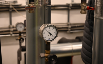 Manometer an einem Rohr, das den Druck anzeigt. Im Hintergrund sind weitere Rohre und ein Blick auf eine weitere Anzeige sichtbar. Die Umgebung deutet auf eine technische Einrichtung oder ein Versorgungssystem hin.