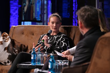 Eine Person mit einem schwarz-weißen Oberteil sitzt auf einem Sofa und spricht in ein Mikrofon. Neben ihr stehen Wasserflaschen. Im Hintergrund sind unscharfe Lichter und andere Personen beim Gespräch zu sehen.