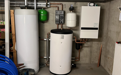 Abbildung eines Heizungsraums mit einer weißen Heizungsanlage, einem Wassertank, einem grünen Expansionsgefäß und verschiedenen Rohren. An der Wand befinden sich auch ein kleiner Wasserbehälter und ein Heizungssteuergerät. Im Vordergrund sind blaue Rohre zu sehen.