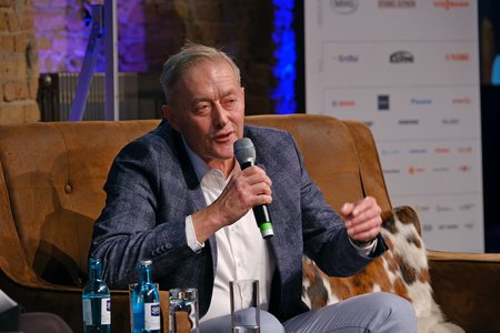 Ein Mann in einem blauen Anzug sitzt auf einem Sofa und spricht in ein Mikrofon. Vor ihm steht ein Glas Wasser und mehrere Wasserflaschen. Der Hintergrund besteht aus gemauerter Wand und beleuchteten Elementen.