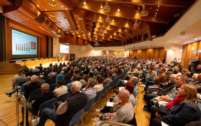 Eine große Menschenmenge sitzt in einem modernen Konferenzsaal. Eine Präsentation wird an eine Leinwand projiziert, während eine Person auf der Bühne spricht. Der Raum hat eine attraktive Holzausstattung und ist gut beleuchtet.