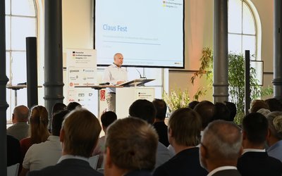 Ein Redner steht an einem Podium und spricht vor einem Publikum in einem hellen Raum mit großen Fenstern. Auf dem Bildschirm und im Hintergrund sind Präsentationsmaterialien zu sehen. Das Publikum, bestehend aus mehreren Personen, hört aufmerksam zu.