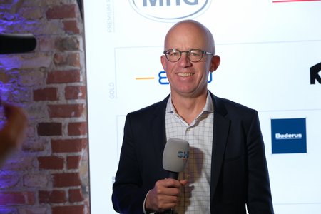 Ein Mann mit Brille steht vor einer Wand mit Logos und Sponsorenbeschriftungen. Er hält ein Mikrofon in der Hand und lächelt. Der Hintergrund zeigt eine Backsteinwand und einige weiße Grafiken.