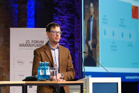 Ein Mann steht hinter einem Rednerpult auf einer Konferenz. Er trägt einen braunen Blazer und hat eine Brille auf. Auf dem Bildschirm hinter ihm sind Informationen zu Wärmepumpen und eine Darstellung von Daten zu sehen. Wasserflaschen stehen auf dem Pult.