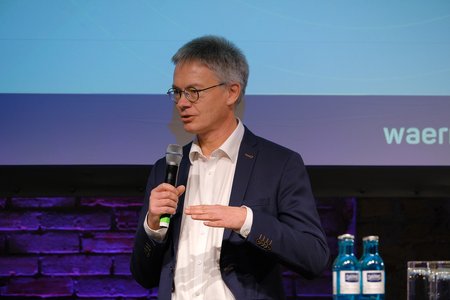 Ein Mann mit Brille und Kurzhaarschnitt steht an einem Mikrofon und spricht. Er trägt einen dunkelblauen Anzug und hat einen weißen Hemd auf. Im Hintergrund sind Wasserflaschen sichtbar. Die Szene vermittelt eine professionelle Präsentationsatmosphäre.