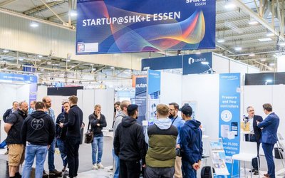 Auf der Messe SHK Essen stehen Menschen zusammen, diskutieren und tauschen Ideen aus. Im Hintergrund ist ein Banner mit der Aufschrift "STARTUP@SHK+E ESSEN" zu sehen. Stände präsentieren verschiedene Produkte und Innovationen.