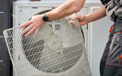 Ein Handwerker entfernt ein Gitter von einer Klimaanlage. Die Person trägt eine Arbeitskleidung und hält das Gitter mit beiden Händen. Im Hintergrund ist die Klimaanlage sichtbar.