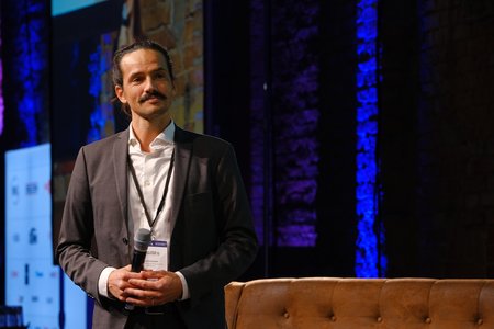 Ein Mann mit Schnurrbart steht auf einer Bühne und hält ein Mikrofon. Er trägt einen Anzug und blickt in die Kamera. Im Hintergrund sind farbige Lichtakzente und eine Leinwand mit Präsentationselementen sichtbar.