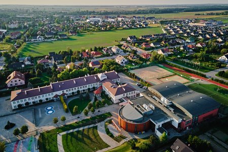 Eine Luftaufnahme eines Wohngebiets mit grünen Feldern und vielen Häusern. Im Vordergrund ist ein Schulgebäude zu sehen, ergänzt durch Sportanlagen und gepflegte Grünflächen. Die Umgebung wirkt ruhig und ländlich.