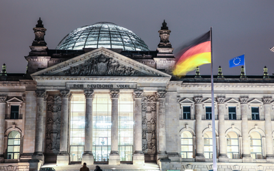 Das Bild zeigt die Reichstagsgebäude in Berlin bei Nacht. Im Vordergrund wehen die deutsche und die europäische Flagge. Die Fassade des Gebäudes ist mit beeindruckenden architektonischen Details verziert, darunter große Fenster und eine Kuppel.