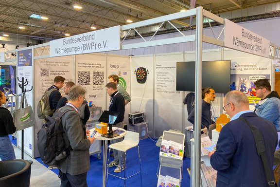Ein Messestand des Bundesverbandes Wärmepumpe (BWP) e.V. ist abgebildet. Mehrere Personen diskutieren und interagieren am Stand, der mit Informationsmaterial und einem Bildschirm ausgestattet ist. Im Hintergrund sind Plakate und QR-Codes zu sehen, die das Thema Wärmepumpen unterstützen.
