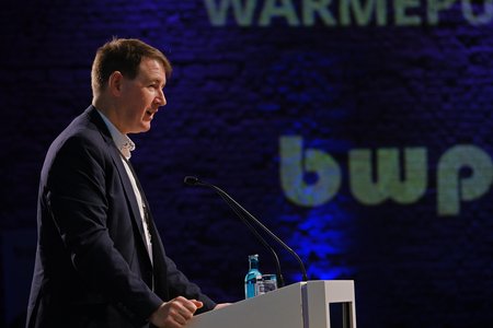 Ein Mann steht an einem Rednerpult und hält eine Präsentation. Im Hintergrund sind verschwommene Logos und Text zu sehen, die auf eine Veranstaltung hinweisen. Der Fokus liegt auf dem Sprecher und seiner Präsentation.