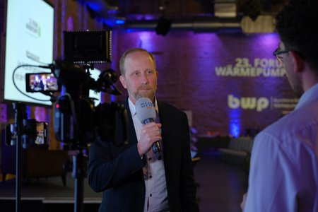 Ein Mann in einem Anzug hält ein Mikrofon und spricht in die Kamera. Im Hintergrund ist eine Veranstaltung mit dem Titel "23. Forum Wärmepumpen" und der Schriftzug “bwp” zu sehen. Die Atmosphäre ist professionell und gut beleuchtet.