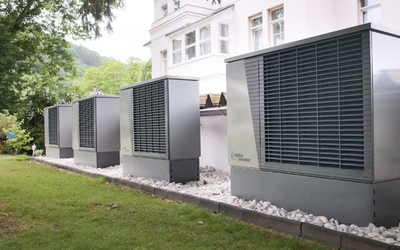 Vier moderne Wärmepumpen stehen auf einem Rasen, nebeneinander entlang einer Hauswand. Im Hintergrund ist ein reproduzierbares, helles Gebäude mit Fenster zu sehen, umgeben von Bäumen. Der Boden ist mit kleinen, weißen Steinen bedeckt.