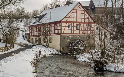 Ein historisches, zweigeschossiges Gebäude mit Fachwerkfassade steht an einem schneebedeckten Ufer. Neben dem Haus fließt ein ruhiger Wasserlauf, in dem ein Wasserrad sichtbar ist. Die Umgebung ist winterlich mit schneebedeckten Bäumen und einem grauen Himmel.