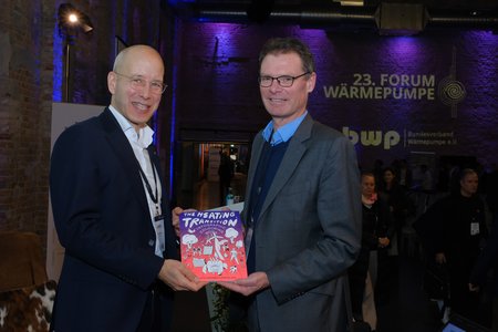 Zwei Männer stehen in einem Veranstaltungsraum und lächeln in die Kamera. Einer von ihnen hält ein Buch mit dem Titel "The Heating Transition" in den Händen. Im Hintergrund sind unscharf Menschen und ein Banner des 23. Forums Wärmepumpe sichtbar.