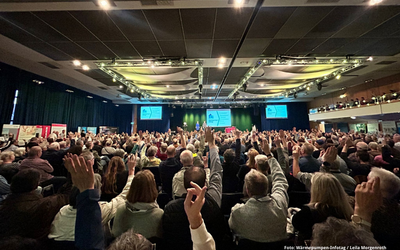 Eine große Menschenmenge in einem Konferenzsaal hebt die Hände. Im Hintergrund sind Bildschirme mit Informationen zu sehen. Die Stimmung ist aktiv und engagiert, was auf eine lebhafte Diskussion oder Abstimmung hindeutet.