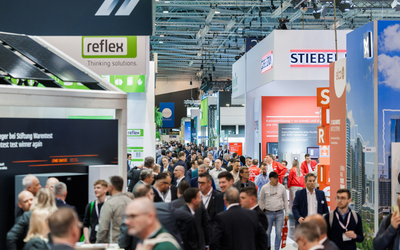 Die Aufnahme zeigt eine belebte Messehalle mit zahlreichen Besuchern, die zwischen verschiedenen Ständen flanieren. Im Hintergrund sind Werbeschilder und Stände von Unternehmen wie "reflex" und "STIEBEL" zu sehen. Die Atmosphäre wirkt geschäftig und informativ.