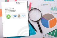 Das Bild zeigt eine Broschüre mit dem Titel "Branchestudie 2026" und den Untertiteln "Marktentwicklung, Prognosen & Handlungsempfehlungen". Neben der Broschüre liegen verschiedene Diagramme und Grafiken, darunter ein Tortendiagramm und Balkendiagramme, sowie eine Lupe und ein Stift.