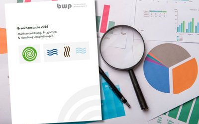 Das Bild zeigt eine Broschüre mit dem Titel "Branchestudie 2026" und den Untertiteln "Marktentwicklung, Prognosen & Handlungsempfehlungen". Neben der Broschüre liegen verschiedene Diagramme und Grafiken, darunter ein Tortendiagramm und Balkendiagramme, sowie eine Lupe und ein Stift.