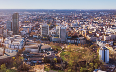 Luftaufnahme einer Stadt, die moderne Hochhäuser und historische Gebäude zeigt. Im Vordergrund sind grüne Parkanlagen sichtbar. Die Skyline erstreckt sich über eine weite Fläche, mit einer klaren Sicht auf die städtische Umgebung und den blauen Himmel.