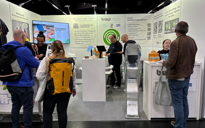 Ein Messestand mit mehreren Personen, die sich um einen Tisch versammeln. Auf dem Tisch befinden sich Geräte und Informationsmaterialien. Im Hintergrund sind Werbematerialien und Grafiken zur Firma "bwp" zu sehen. Die Atmosphäre ist lebhaft und informativ.