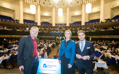 Drei Personen stehen auf einer Bühne in einem großen Veranstaltungsraum. Der Hintergrund zeigt eine große Menge von Zuhörern an Tischen. Ein Schild mit der Aufschrift "Wärmepumpen Initiative" ist sichtbar. Die Atmosphäre vermittelt eine formelle Veranstaltung oder Konferenz.