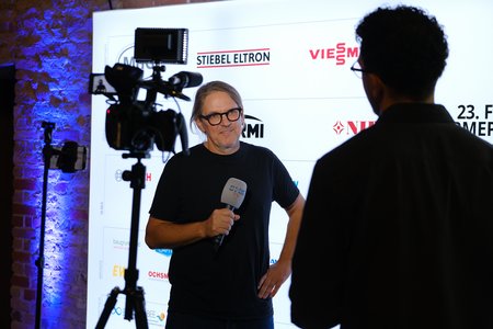 Eine Person mit Brille steht im Vordergrund und hält ein Mikrofon. Sie spricht anscheinend mit einem anderen Menschen, der im Schatten steht. Im Hintergrund befinden sich Logos von Sponsoren oder Partnern der Veranstaltung. Es scheint sich um ein Interview oder eine Präsentation zu handeln.