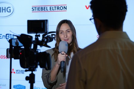 Eine Frau spricht in ein Mikrofon vor einer Kameralinse. Im Hintergrund sind Logos verschiedener Unternehmen sichtbar. Die Szene scheint ein Interview oder eine Pressekonferenz zu sein, wobei die Frau fokussiert und professionell wirkt.