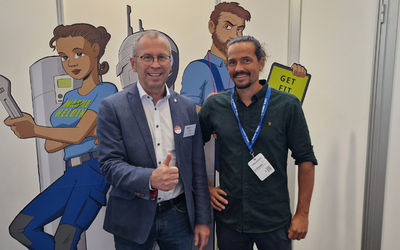 Zwei Erwachsene stehen vorne vor einer Wand mit Comicfiguren. Der Mann links trägt einen Anzug und zeigt Daumen hoch, während der Mann rechts in einem dunklen Hemd lächelt. Im Hintergrund sind farbige Zeichnungen von Charakteren zu sehen, die in einem technischen Umfeld arbeiten.