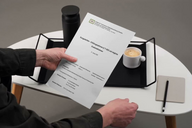 Eine Hand hält ein Dokument mit dem Titel "Eingangs- und Bewerbungsformular". Auf einem Tisch steht eine schwarze Thermoskanne und eine Tasse mit Kaffee. Das Bild vermittelt eine ruhige Arbeitsumgebung.