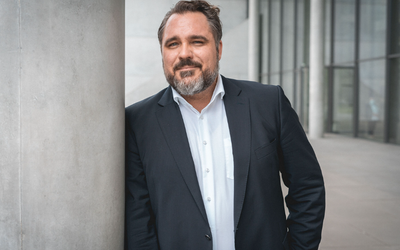 Ein Mann mit Bart trägt einen dunklen Anzug und steht entspannt an einem Säulenstütze. Er hat kurze, leicht gewellte Haare und blickt direkt in die Kamera, wobei er lächelt. Der Hintergrund zeigt eine moderne Architektur.