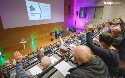 Eine Veranstaltung in einem großen Saal mit zahlreichen Teilnehmenden. Auf der Bühne stehen Personen, die Informationen zu einem Thema präsentieren. Viele Zuhörer heben die Hände, um Fragen zu stellen oder ihre Zustimmung zu zeigen. Im Hintergrund ist eine Präsentationsfolie sichtbar.