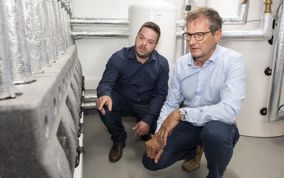 Zwei Männer knien in einem Technikraum. Einer zeigt auf eine technische Vorrichtung, während der andere aufmerksam zuhört. Im Hintergrund sind Rohrleitungen und ein Wasserbehälter sichtbar. Beide Männer tragen formelle Kleidung und scheinen eine fachliche Diskussion zu führen.