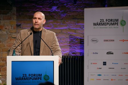 Ein Mann spricht an einem Rednerpult während des 23. Forums Wärmepumpe in Berlin. Im Hintergrund sind Veranstaltungsdetails und Sponsorenlogos zu sehen. Der Mann trägt einen braunen Blazer und hat eine klare, aufmerksame Miene.