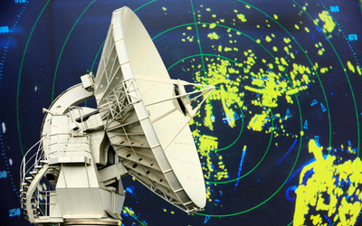 Ein großer Satellitenempfänger steht im Vordergrund, während im Hintergrund ein Radarbild mit gelben und blauen Punkten auf grünem Grund zu sehen ist. Die Grafik zeigt verschiedene Objekte oder Bewegungen, die vom Radar erfasst werden.