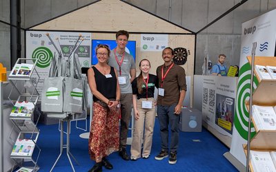 Eine Gruppe von vier Personen steht im Vordergrund eines Messestands. Im Hintergrund sind Informationsmaterialien und Banner zu sehen. Die Personen lächeln und tragen Namensschilder. Die Szenerie vermittelt eine freundliche und einladende Atmosphäre.