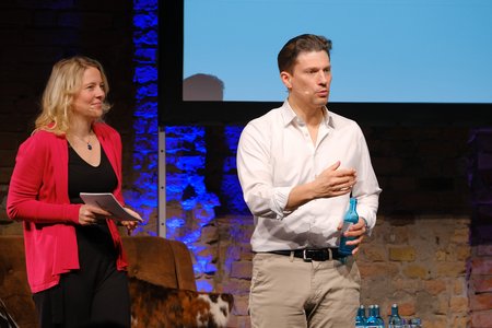 Zwei Personen stehen auf einer Bühne. Eine Frau mit blonden Haaren und einer roten Jacke hält ein Notizblatt, während ein Mann in einem weißen Hemd und beigen Hosen eine Flasche in der Hand hält. Im Hintergrund sind einige Wasserflaschen sichtbar.