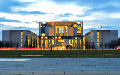 Das Bild zeigt ein modernes Gebäude mit einer beeindruckenden Architektur, beleuchtet in den Abendstunden. Die Fassade besteht aus einem Mix aus Beton und Glas. Im Vordergrund ist ein Platz mit Pflastersteinen und einige grüne Flächen zu sehen.