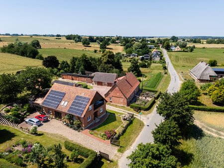 Luftaufnahme eines ländlichen Wohngebiets mit mehreren Häusern. Im Vordergrund steht ein Haus mit Solarpanelen auf dem Dach, umgeben von einem gepflegten Garten. Eine ruhige Straße verläuft entlang der Nachbarhäuser und Wiesen, die die Landschaft dominieren.