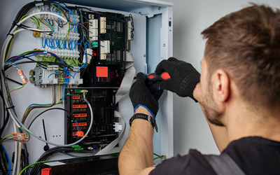 Ein Techniker arbeitet an einem Schaltschrank. Er trägt schwarze Handschuhe und benutzt einen Schraubendreher, um Kabel und Komponenten in einem elektrischen Verteiler zu überprüfen oder zu reparieren. Der Bereich ist mit verschiedenen Drähten und Geräten voll sichtbar.
