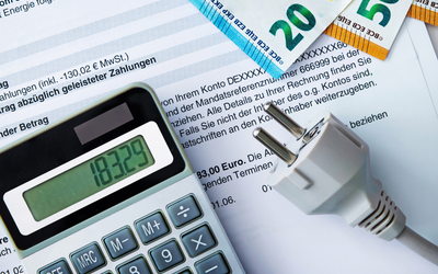 Ein Taschenrechner zeigt den Wert 183,29 an. Daneben liegt ein Stromstecker, und im Hintergrund sind Geldscheine (20 und 50 Euro) sowie ein Dokument mit Informationen zu Zahlungen zu sehen.