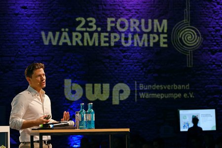 Ein Mann spricht auf einer Bühne beim 23. Forum Wärmepumpe. Im Hintergrund sind das Logo des Bundesverbands Wärmepumpe e.V. und eine stylisierte Darstellung eines Wärmepumpensystems sichtbar. Auf dem Tisch stehen mehrere Wasserflaschen.