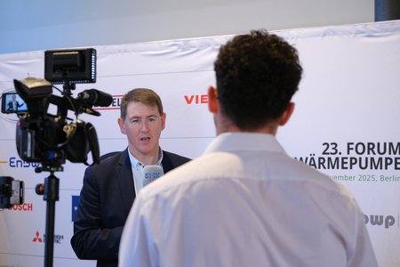 Ein Mann spricht in ein Mikrofon vor einer Kamera. Er trägt einen Anzug und informiert über das 23. Forum Wärmepumpen, das im November 2025 in Berlin stattfindet. Im Hintergrund sind Logos von Sponsoren und Veranstaltern sichtbar.